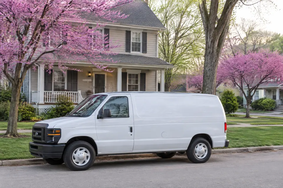 A white service van parked outside a two-story home on a street lined with blooming redbuds